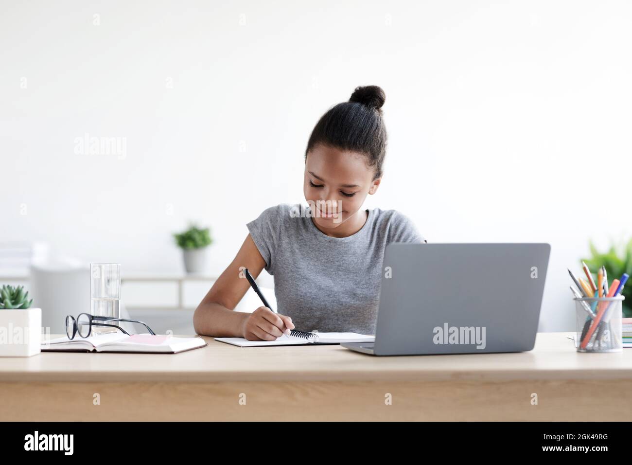Lächelnde junge afroamerikanische Schülerin schreibt im Notizbuch am Computer, studiert Stockfoto
