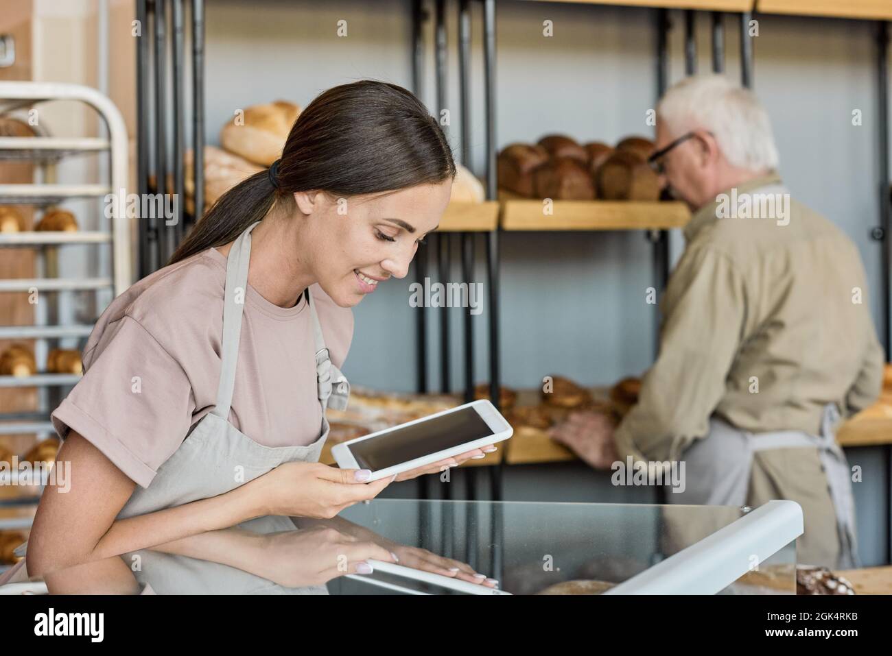 Lächelnde junge Bäckerin auf der Schürze, die mit einem Tisch gegen ihren reifen männlichen Kollegen auflegte, der frisches Brot auf das Regal stellte Stockfoto