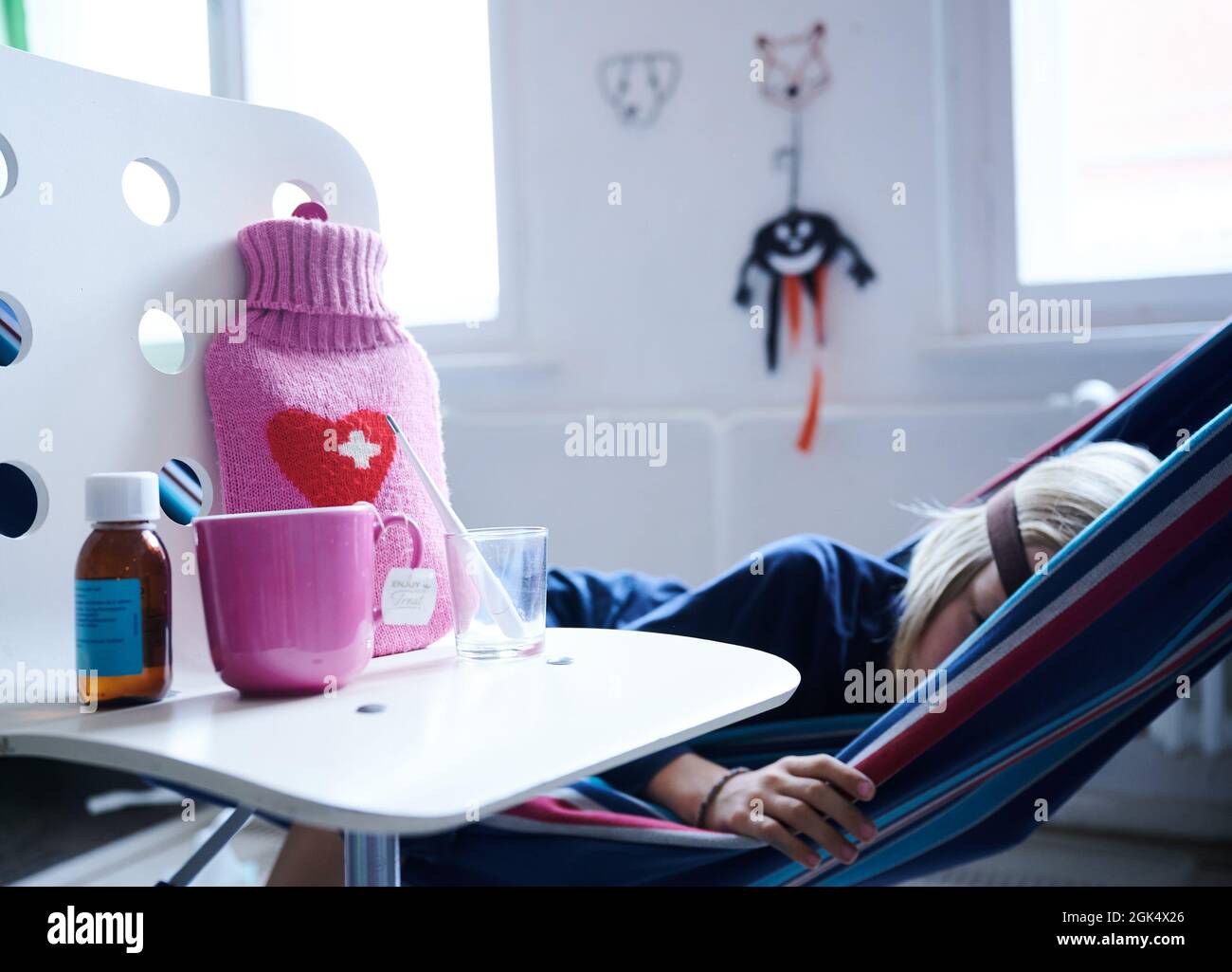 Berlin, Deutschland. September 2021. Ein Fieberthermometer, Hustensirup, eine Heißwasserflasche und eine Tasse Tee stehen auf einem Stuhl, während ein Kind in einer Hängematte im Hintergrund liegt. Quelle: Annette Riedl/dpa/Alamy Live News Stockfoto