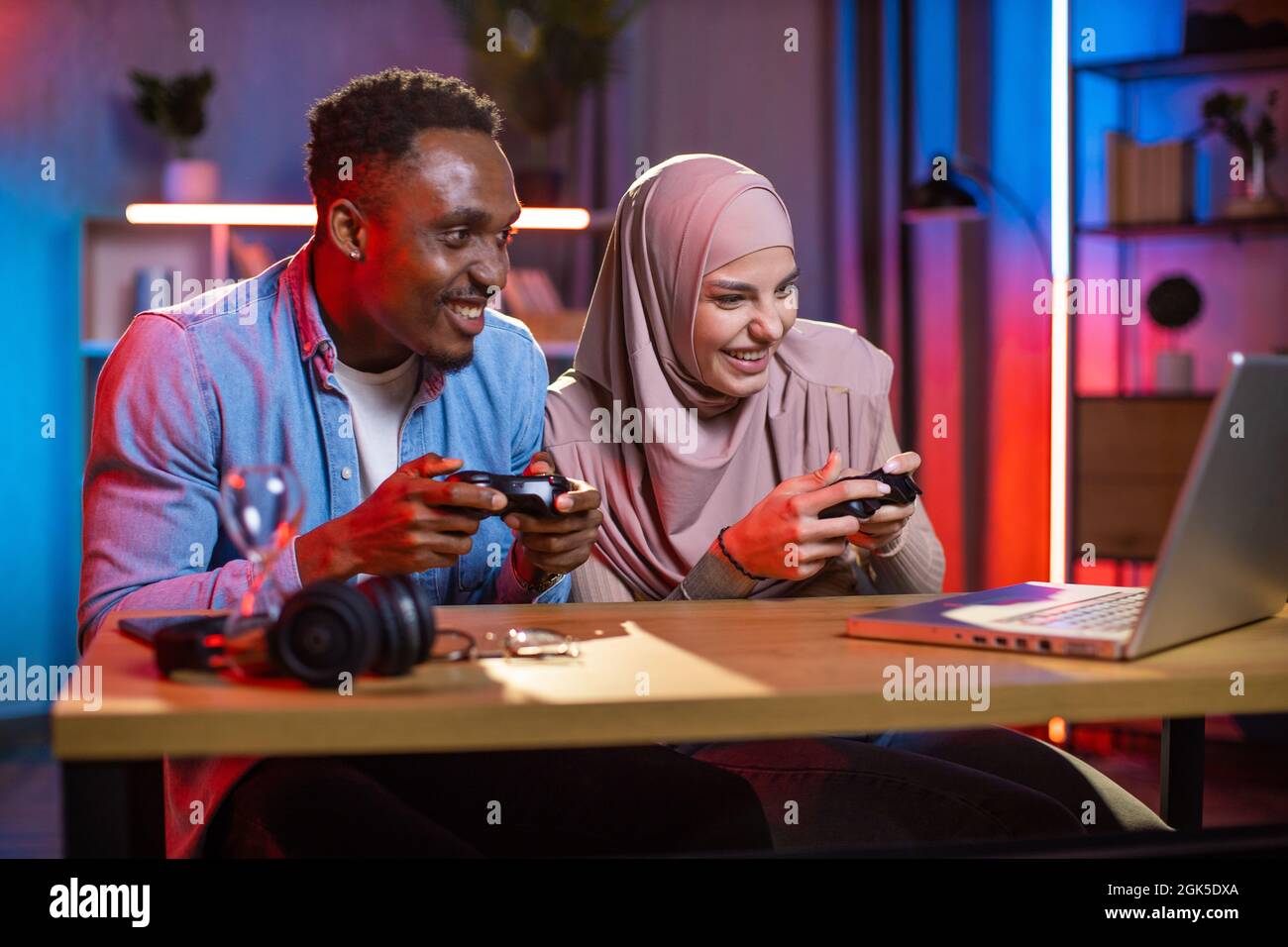 Aufgeregt multiethnischen Paar mit Joysticks in den Händen Blick auf den Computerbildschirm, während Videospiele spielen. Konkurrenzmodus des afrikanischen Mannes und der muslimischen Frau. Häusliches Leben mit Spaß. Stockfoto