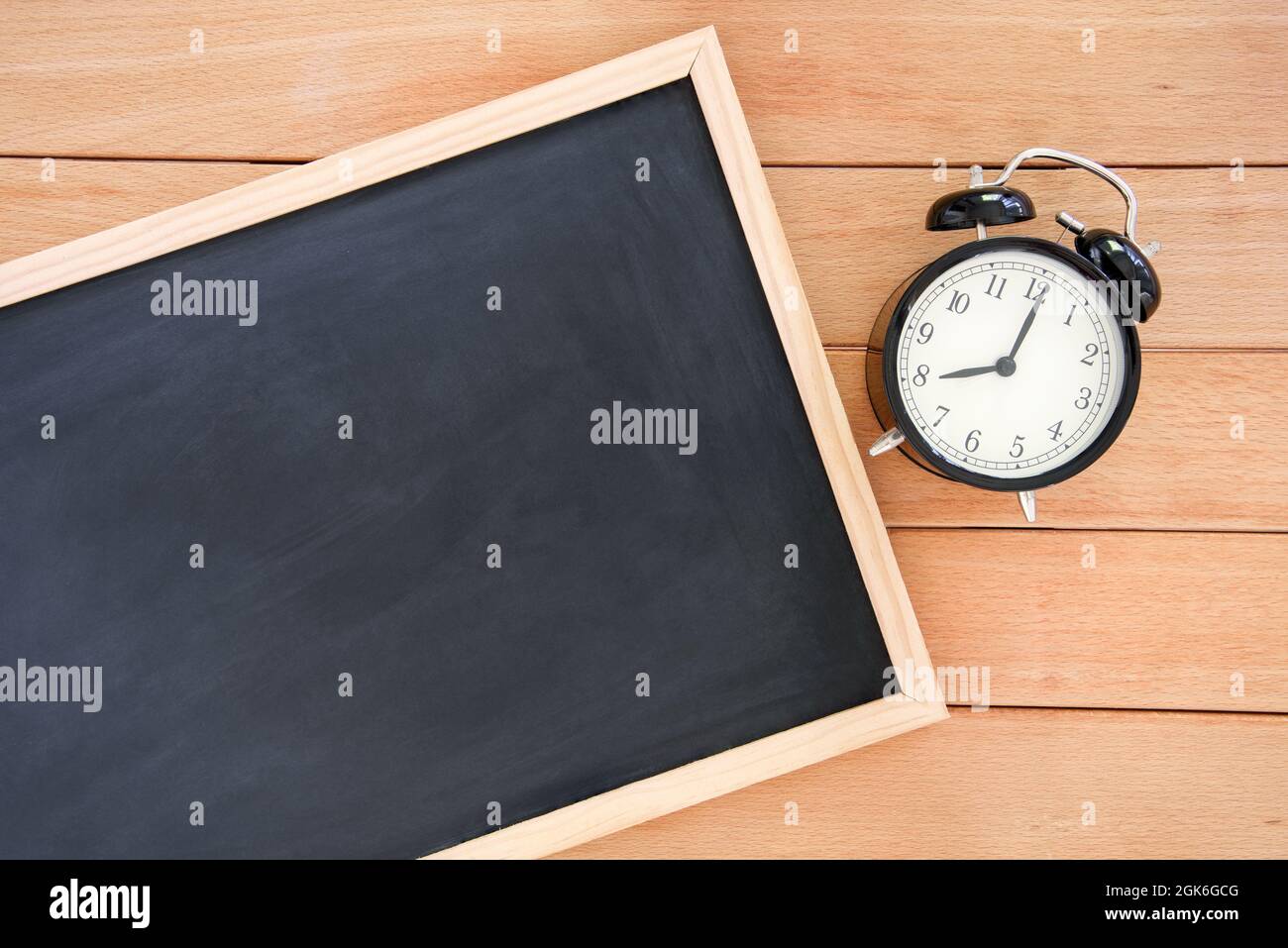 Leere kleine Tafel, die auf einem Holzschreibtisch in der Nähe eines Weckers liegt. Platz für Text. Zurück zum Schulkonzept. Stockfoto