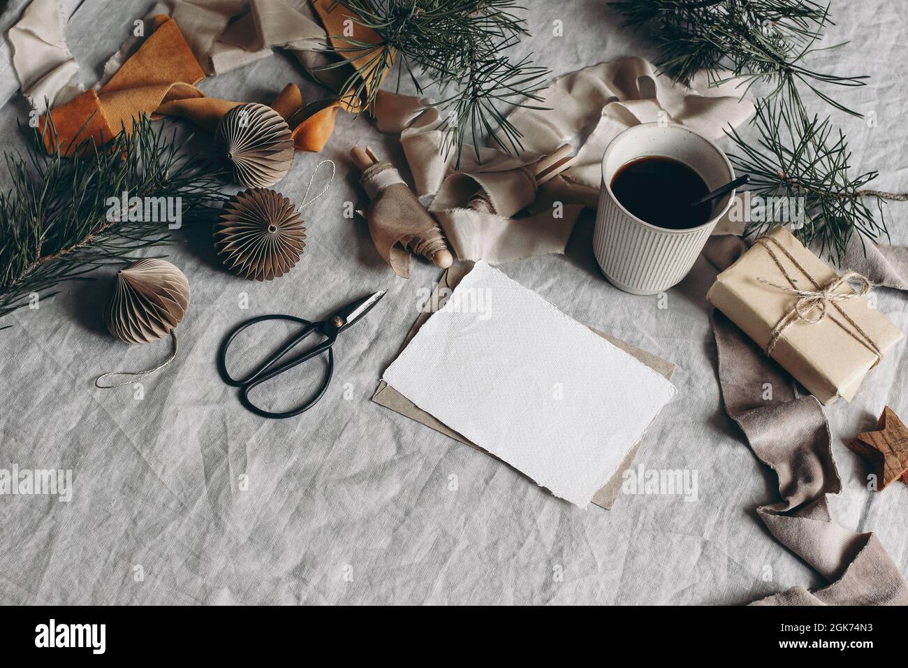 Stillleben zu Weihnachten. Modell mit leerer Grußkarte. Tasse Kaffee, Papierschmuck und Geschenkbox. Samtbänder, Kiefernzweige mit Schere. Leinen Stockfoto