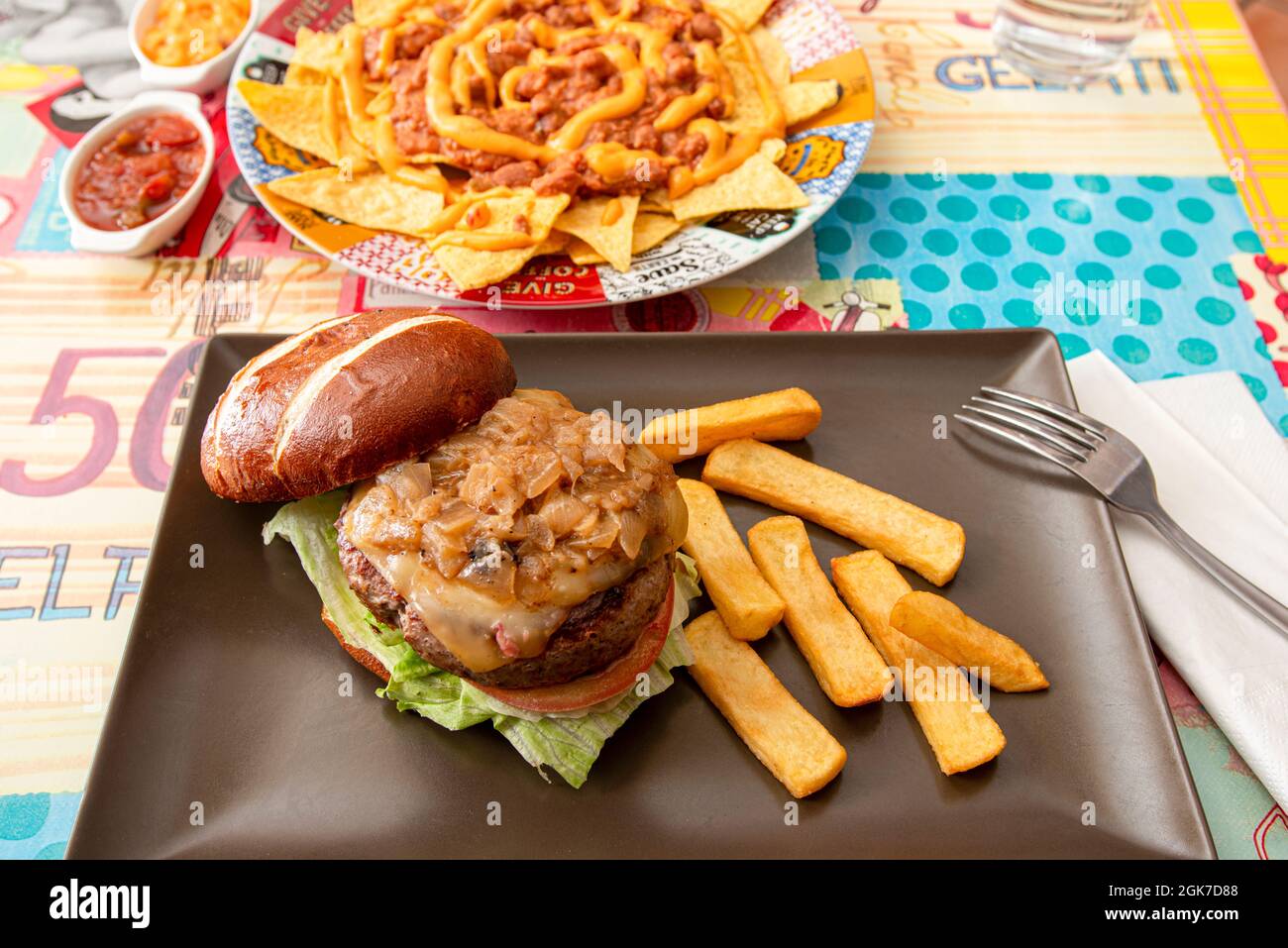 Rindfleisch-Burger mit vielen geschmorten Zwiebeln, Toast, Eisbergsalat, geschmolzenem Käse und Tomaten, serviert mit pommes frites an einem Restauranttisch mit Chili con Stockfoto