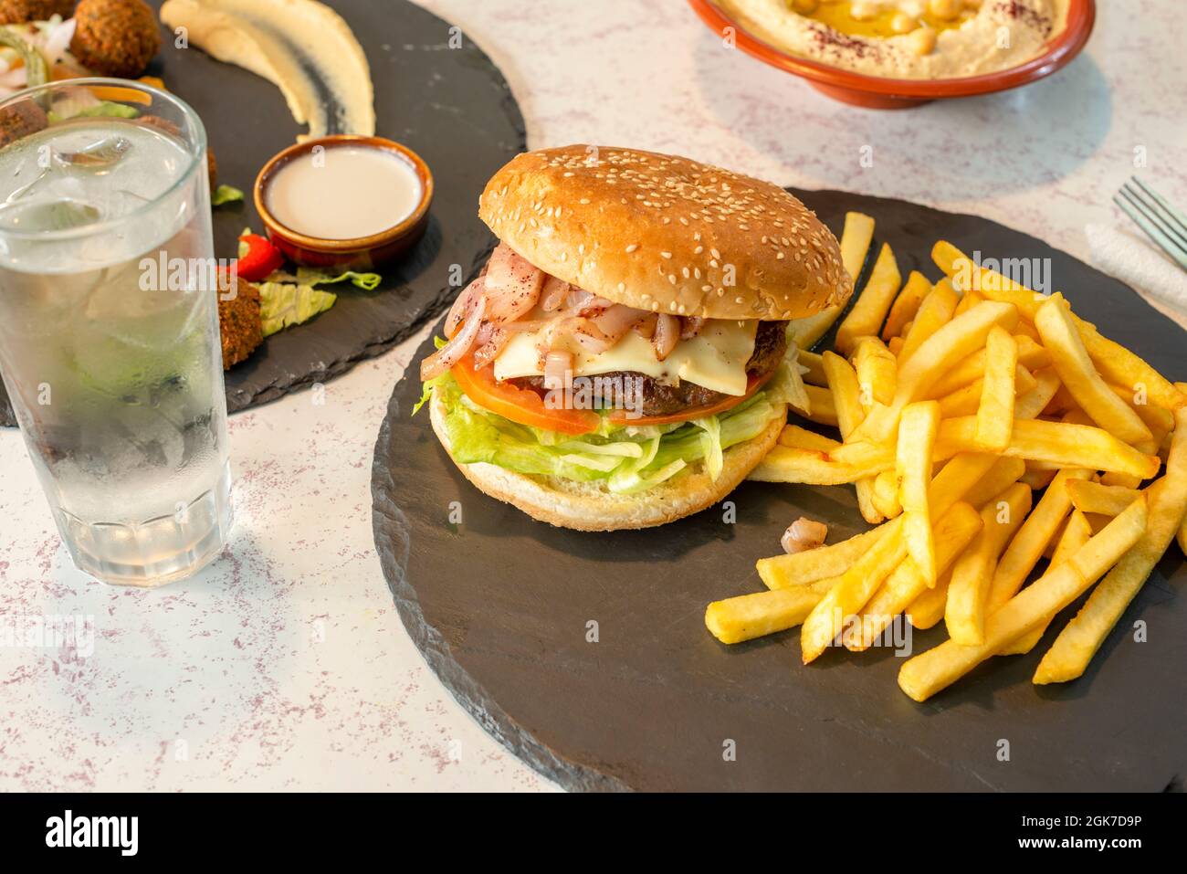Klassischer Rindfleischburger mit Käse und gehackten Zwiebeln, pommes Frites und einem Glas Eiswasser auf einem runden Schieferteller Stockfoto