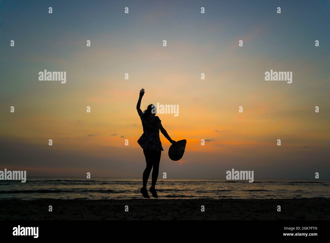 Rückansicht einer isolierten jungen Frau, die in der Luft springt, einen großen Sonnenhut in der Hand hält, bei Sonnenuntergang an einem britischen Strand, während eines Sommerurlaubs. Springen Sie vor Freude. Stockfoto