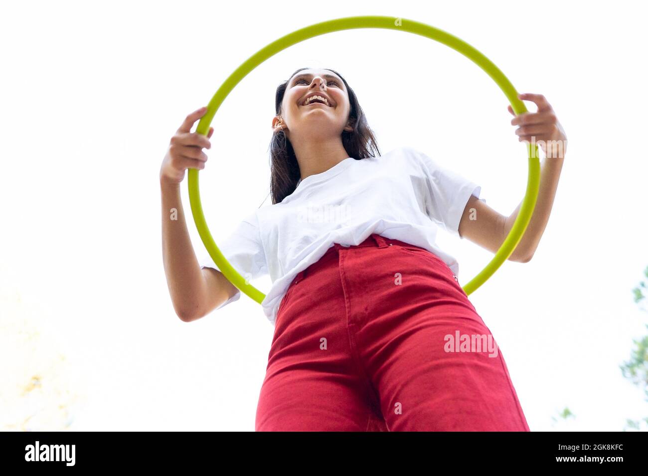 Von unten fröhliche weibliche Teenager in roten Jeans wirbelnden Hula Hoop Reifen während der Freizeit im Park Stockfoto
