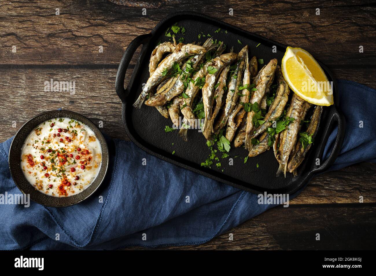 Draufsicht auf leckere gebratene Sardellen mit saftigem Zitronenstück und gehackter Petersilie auf Tablett gegen herzhafte Sauce Stockfoto