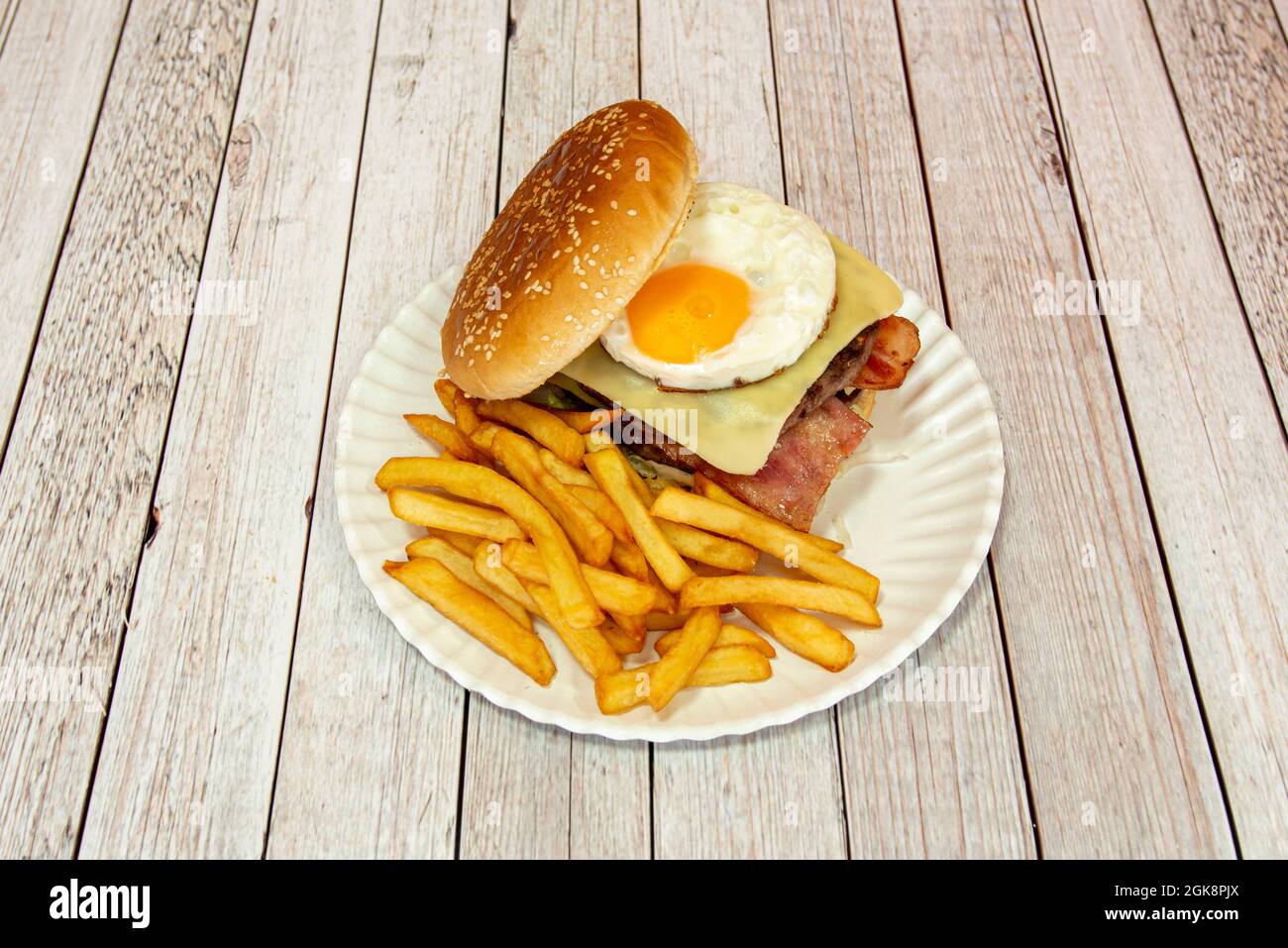 Rinderburger mit Spiegelei, Käse in Scheiben, gebratenem Speck, Gurken und Salat mit einer Portion Pommes auf weißem Teller Stockfoto