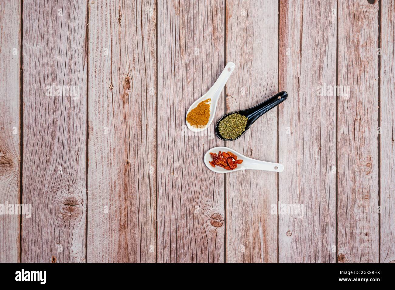 Drei Porzellanlöffel, einer schwarz und zwei weiß mit ganzen Cayenne, gelbem, gelbem, getrockneten Oranganoblättern auf einem Holztisch Stockfoto