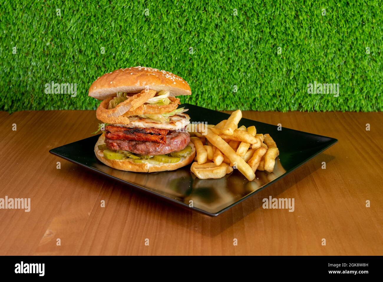 Gemischter Doppel-Burger mit Rind- und Hühnchen, eingelegten Pickle-Scheiben, gebratenen panierten Zwiebelringen, getrockneter Tomate, Eisbergsalat und Pommes Stockfoto