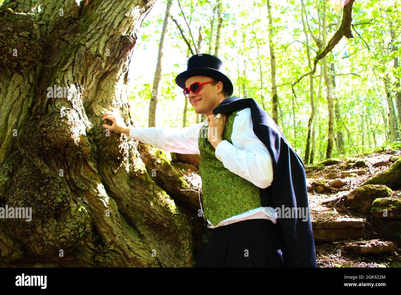 Junger Mann in elegantem Anzug und Hut, der sich im Wald an einem Baum lehnt Stockfoto