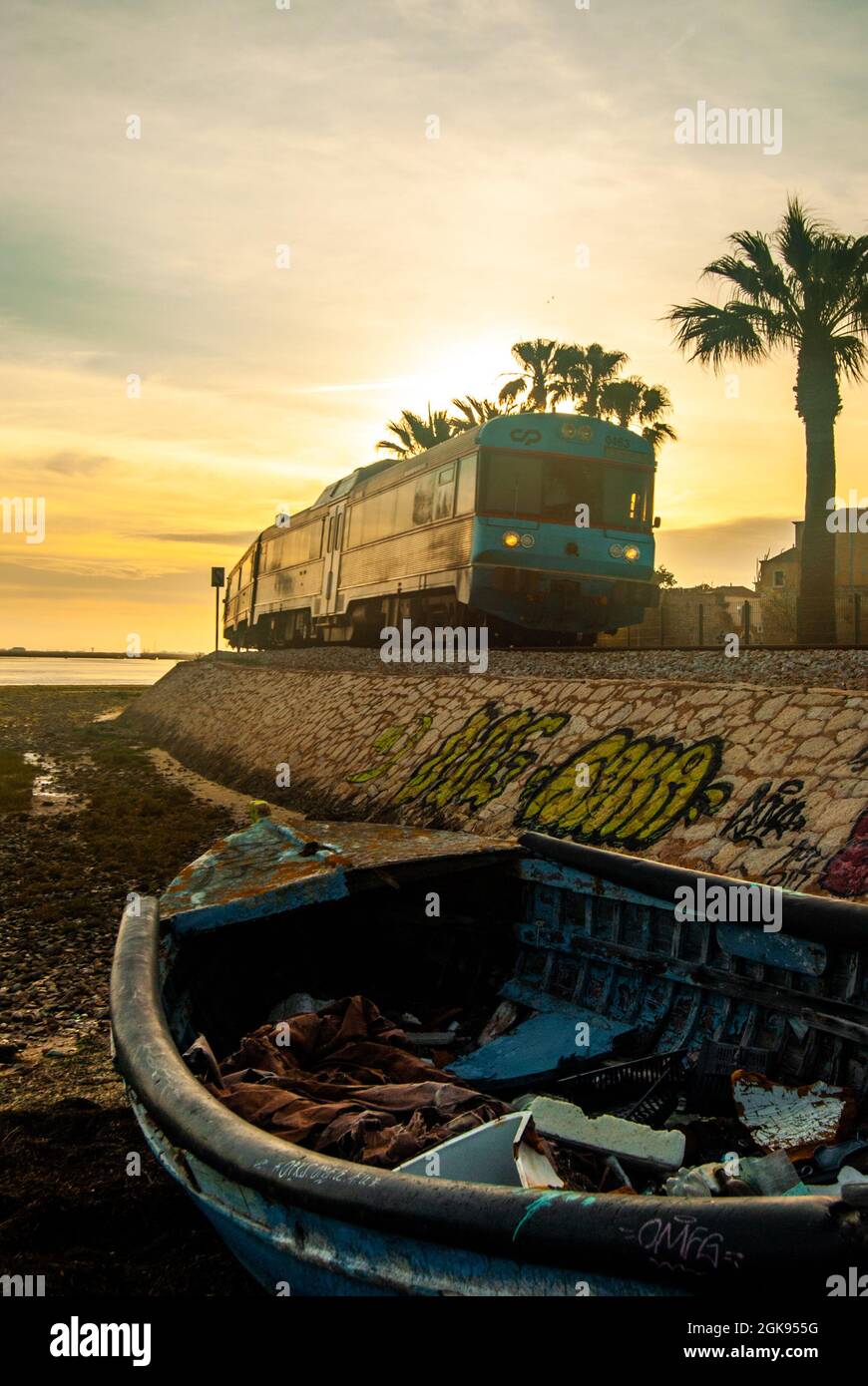 Faro, Portugal - 12. April 2021: Altes Boot am Strand, Pastel-Sonnenuntergang in Faro als ein Zug vorbeifährt, Palmen und die Sonne dahinter Stockfoto