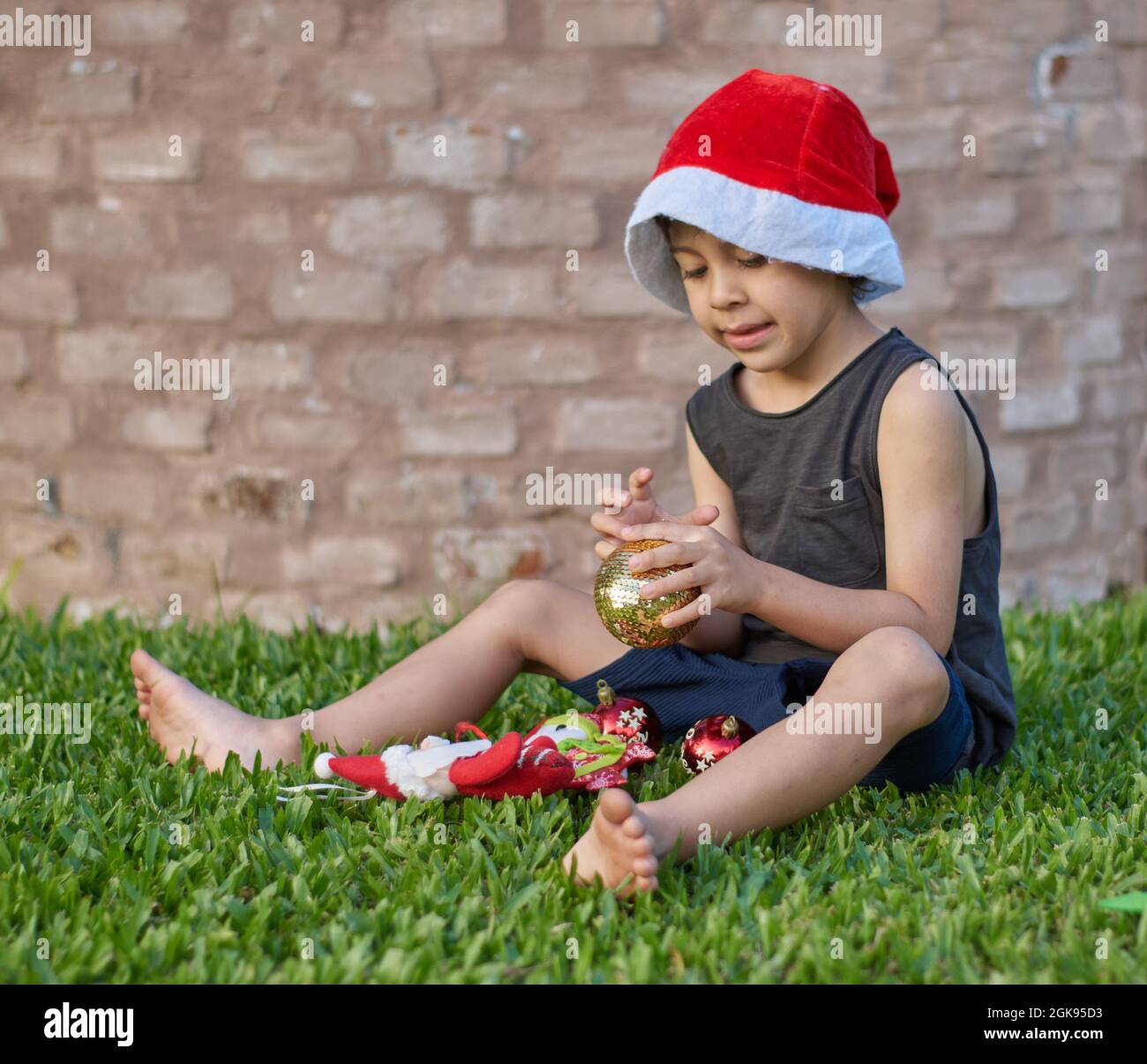 Brauner Junge, der mit weihnachtsschmuck spielt und einen weihnachtsmann-Hut trägt, der auf dem Rasen mit einer Ziegelwand im Hintergrund sitzt. Sommer. Vertikal Stockfoto