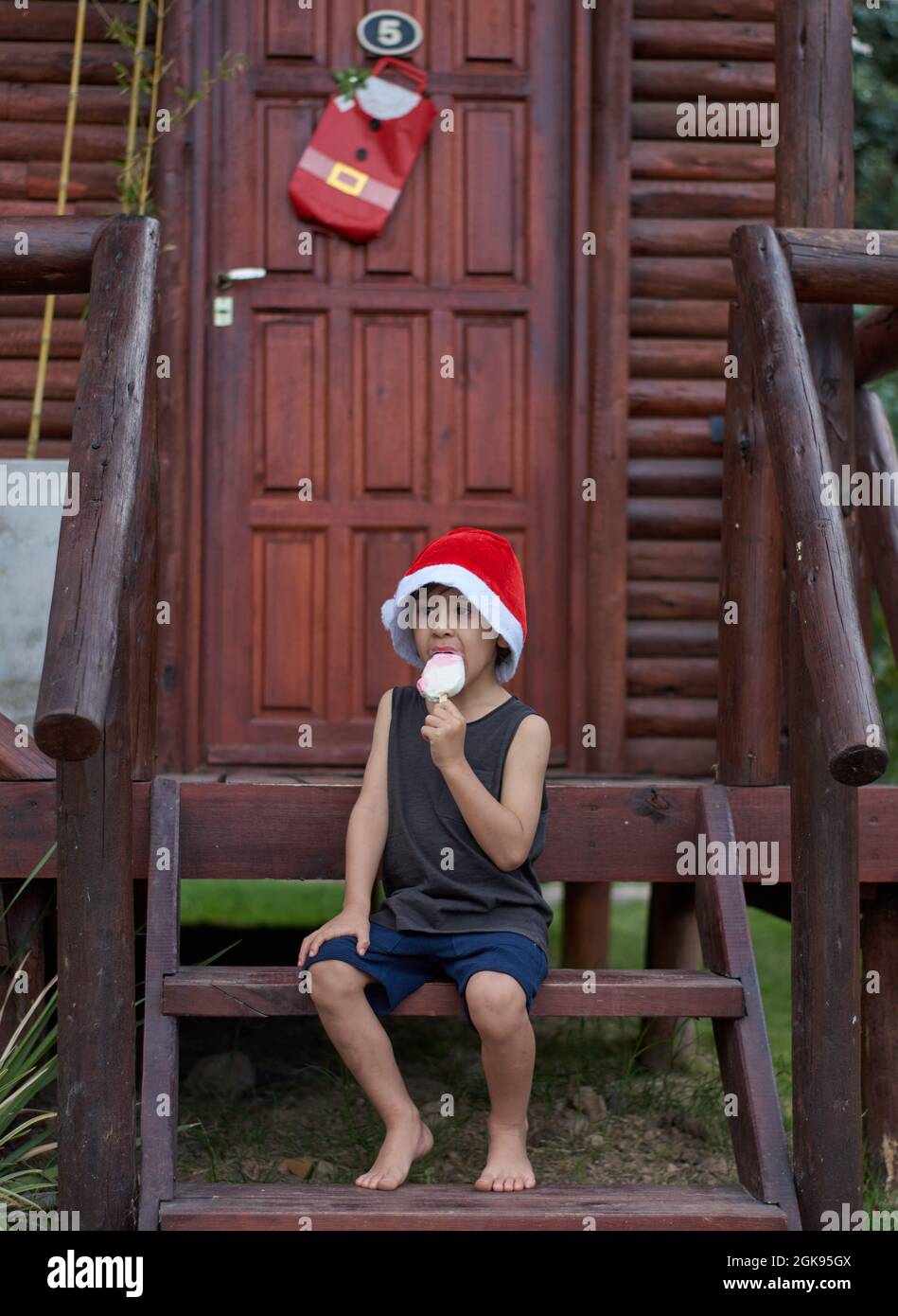 Lateinischer Junge, der zur Weihnachtszeit ein Eis in einem Weihnachtsmannhut genießt und auf der Treppe vor einer braunen Hütte sitzt. Vertikal, Sommer Stockfoto