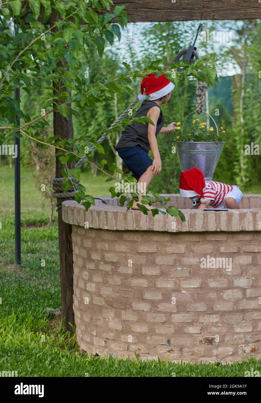 Zwei Kinder mit Weihnachtsmannmützen spielen auf einem geschlossenen Ziegelwasserbrunnen zur Weihnachtszeit. Grüne Vegetation im Hintergrund. Sommer. Vertikal Stockfoto