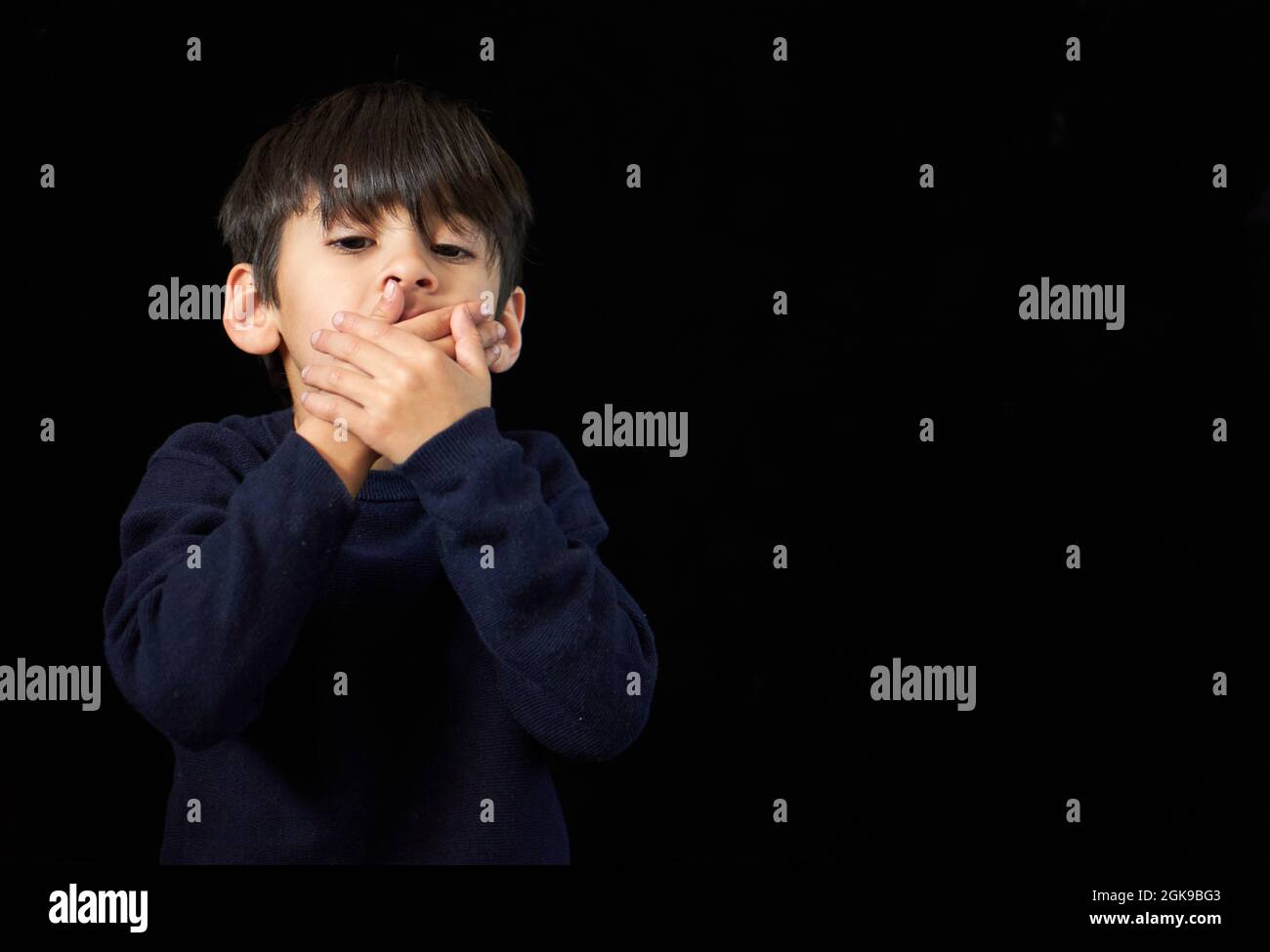 Serious Brunette kleinen Jungen Blick auf die Kamera, die seinen Mund mit beiden Händen. Schwarzer Hintergrund und Kopierraum. Konzept der Stille oder Geheimnis. H Stockfoto
