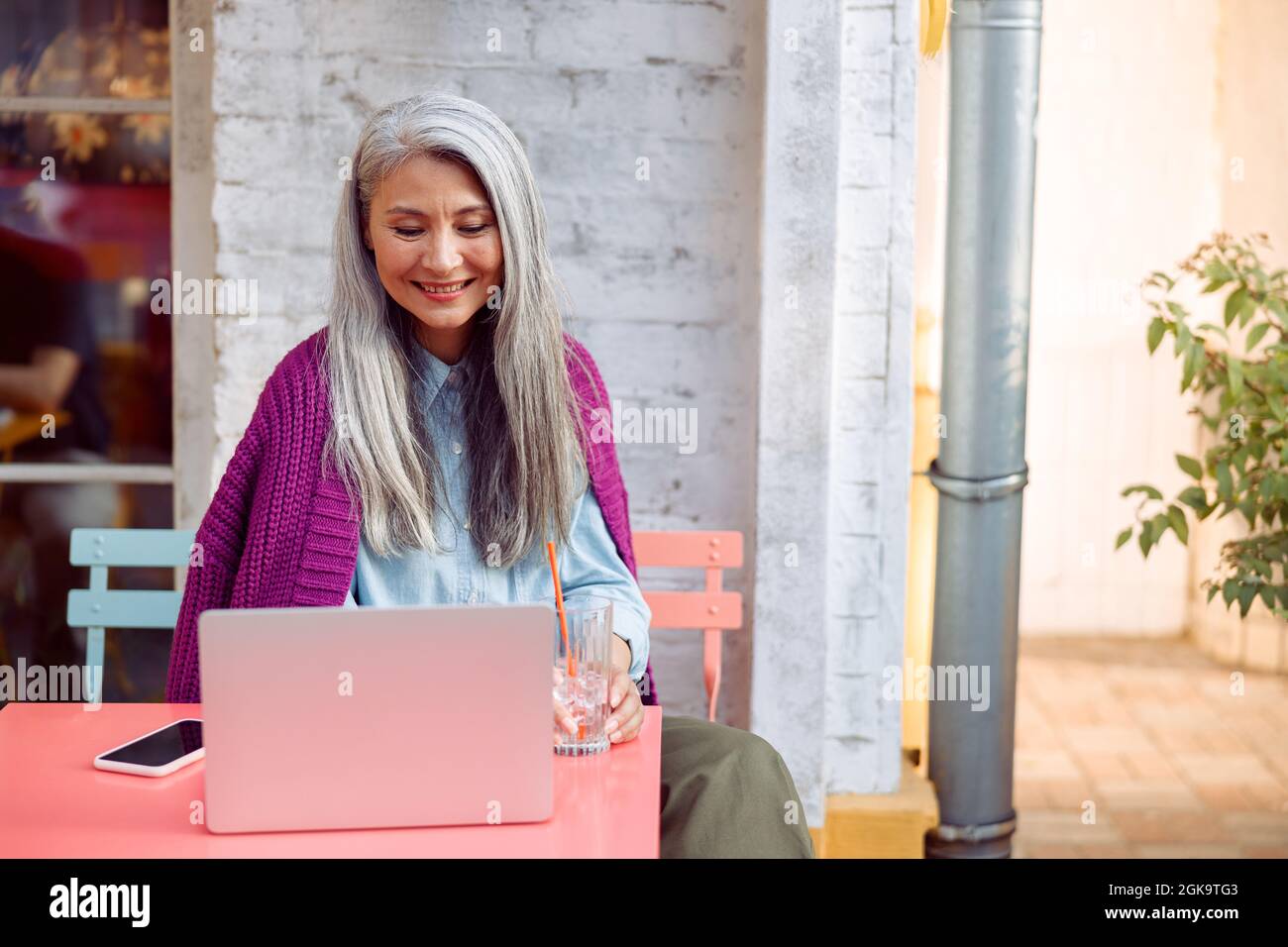 Positive ältere Frau mit Glas und Smartphone schaut auf den Laptop-Bildschirm auf der Terrasse des Cafés im Freien Stockfoto
