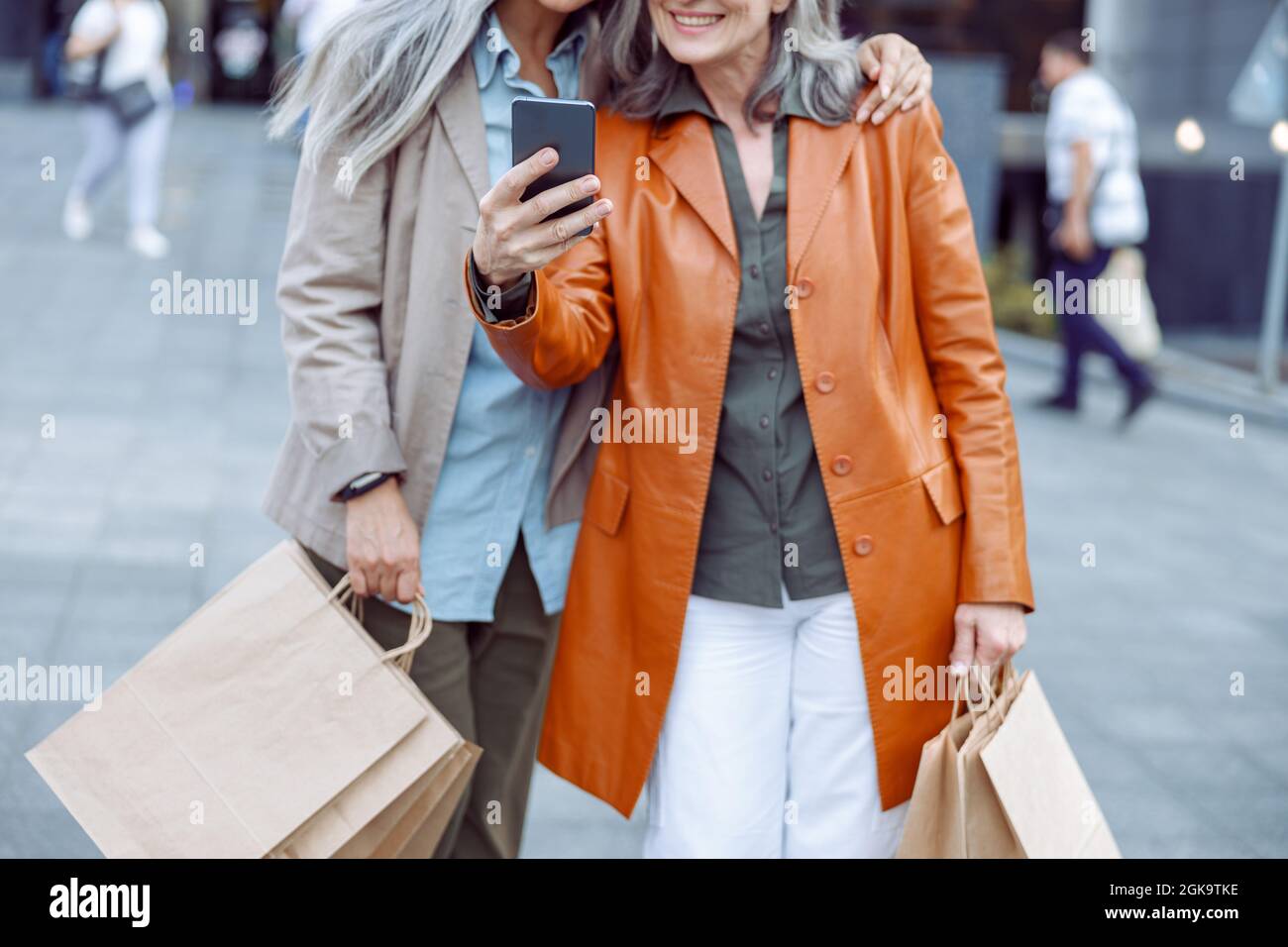 Ältere Dame in Lederjacke und Begleiter mit Einkaufen machen Selfie auf der modernen Stadtstraße Stockfoto