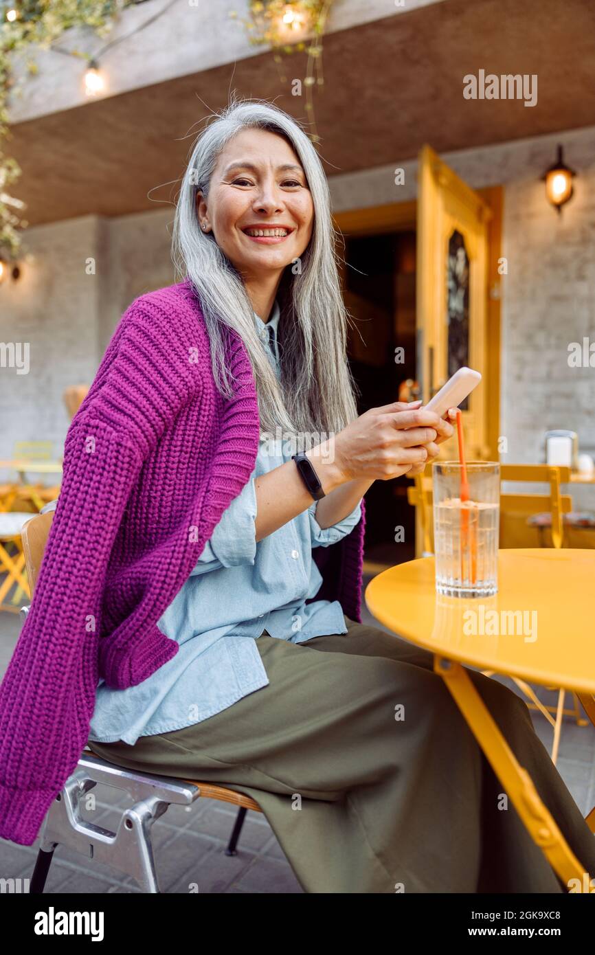 Glückliche ältere asiatische Frau in lila Jacke hält Handy sitzen an kleinen gelben Tisch Stockfoto