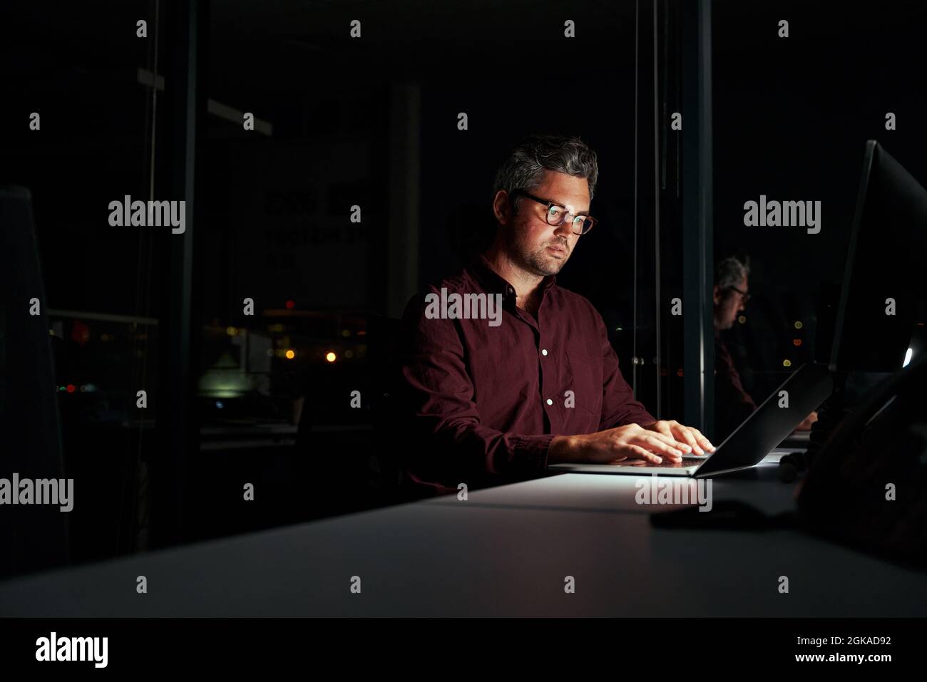 Konzentrierter Geschäftsmann, der spät in der Nacht mit einem Laptop in einem dunklen Büro in der Nähe der französischen Fenster sitzt Stockfoto