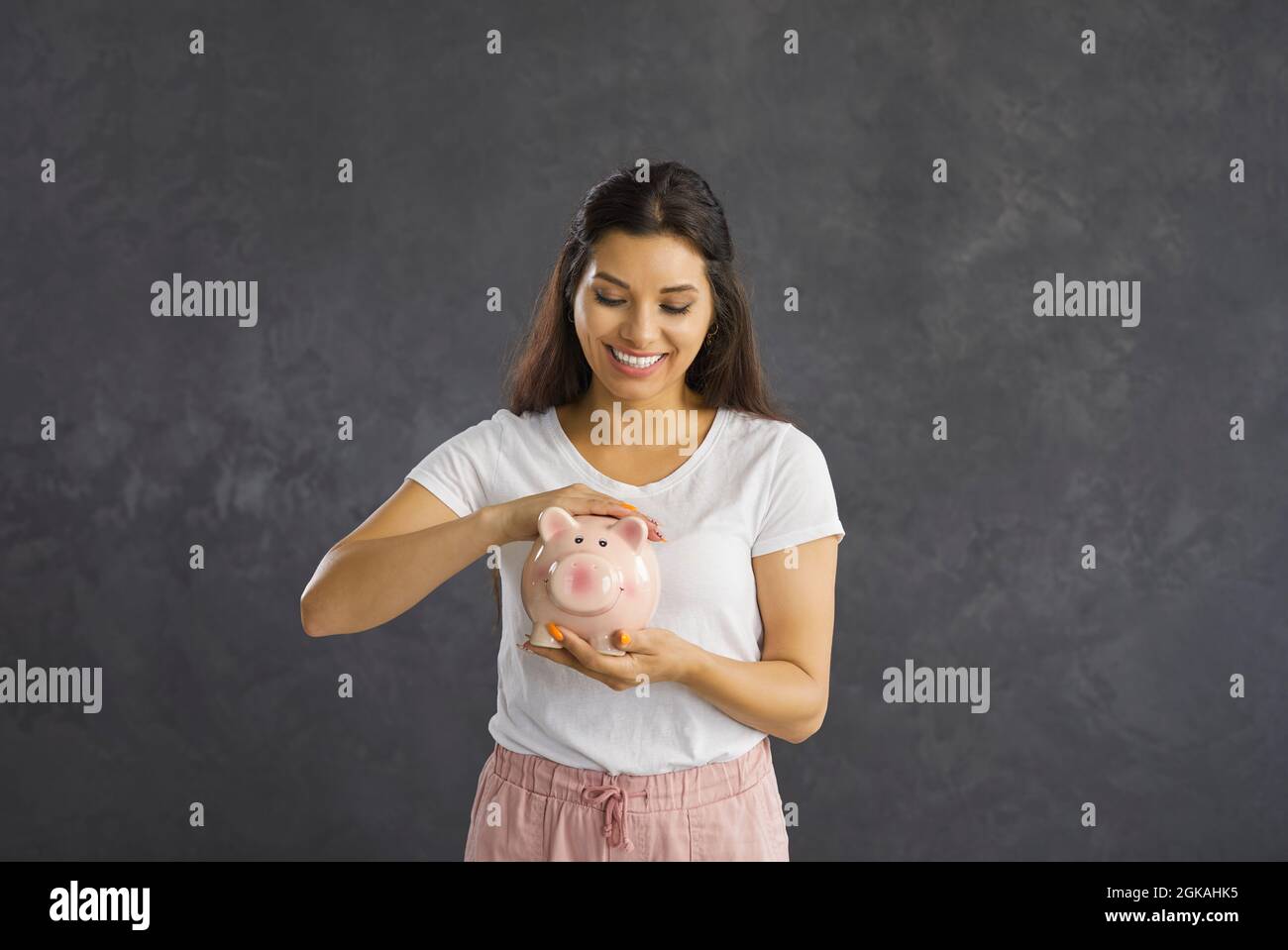 Lächelnde Frau steht auf einem grauen Hintergrund und streichelt liebevoll das Sparschwein mit ihren Ersparnissen. Stockfoto