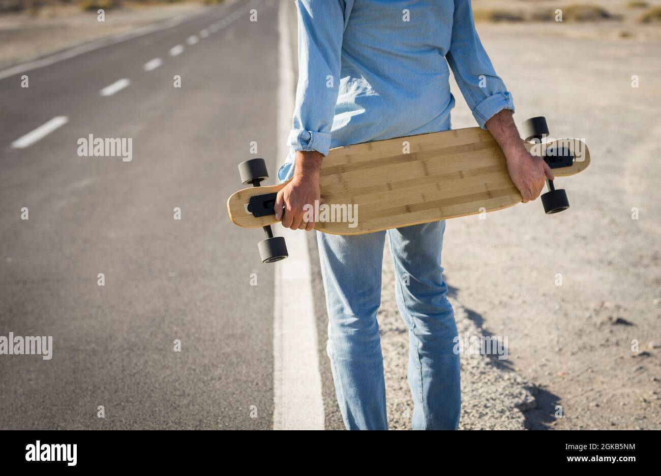 Hände eines Mannes, der Skateboard auf der Straße hält. Rückansicht eines Mannes in Denim-Kleidung mit Skateboard auf einer Landstraße. Sportler mit Skateboard und wai Stockfoto