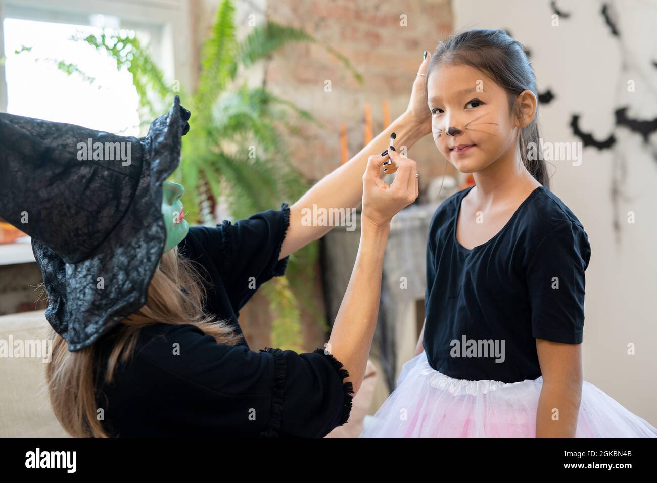 Junge Frau in Hexenhut und schwarzem Pullover Anwendung halloween Make-up auf das Gesicht des niedlichen asiatischen Mädchen vor der Kamera Stockfoto