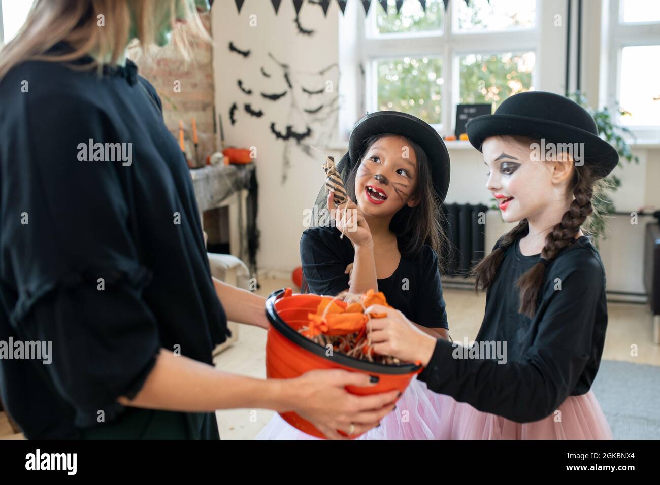 Zwei Mädchen mit bemalten Gesichtern bitten eine junge Frau in schwarzer Kleidung um Halloween-Leckereien Stockfoto
