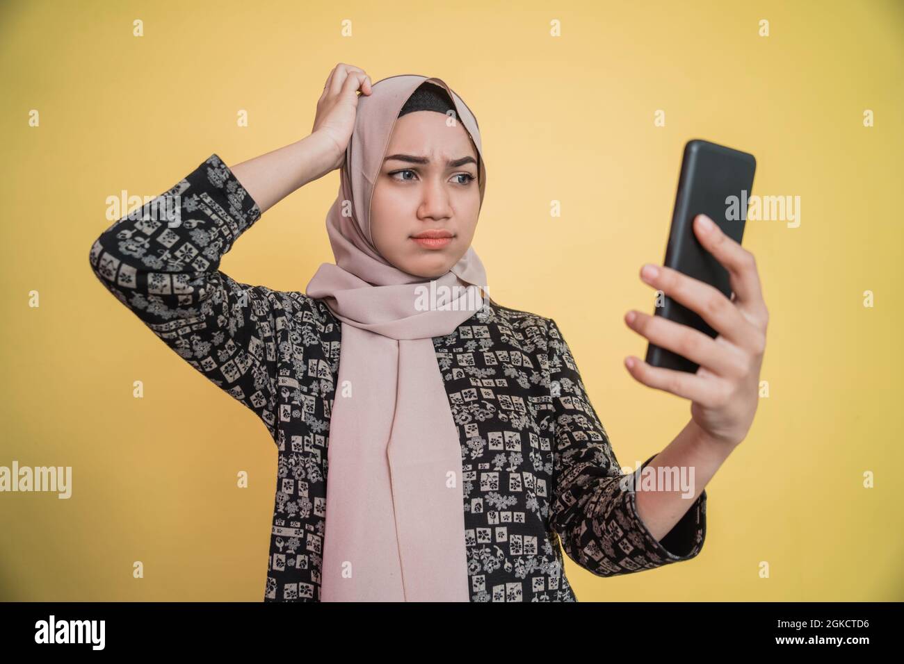 Junge Frau im Hijab verwirrt, während sie auf den Smartphone-Bildschirm mit kratzender Hand schaute Stockfoto