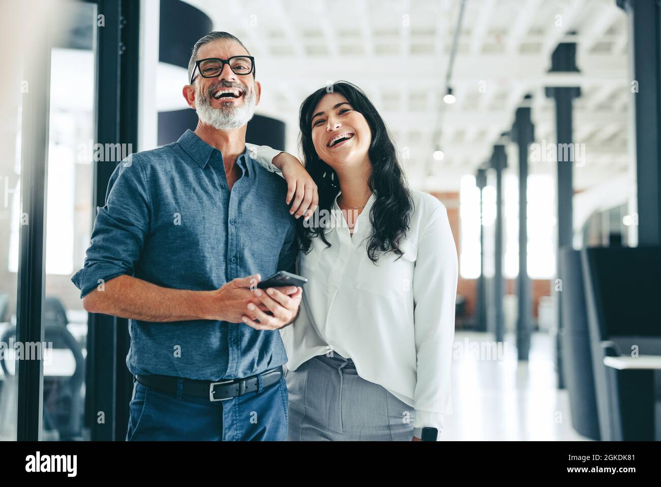 Zwei glückliche Kollegen lächeln fröhlich in einem Büro. Glücklicher, reifer Geschäftsmann, der ein Smartphone hält, während er mit seiner Kollegin in einem Modus steht Stockfoto