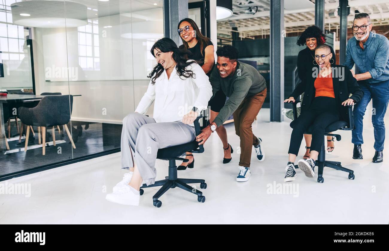 Geschäftsleute, die im Büro Spaß haben. Fröhliche Gruppe von Kollegen, die sich an einem modernen Arbeitsplatz auf Stühlen schieben. Geschäftsteam Stockfoto