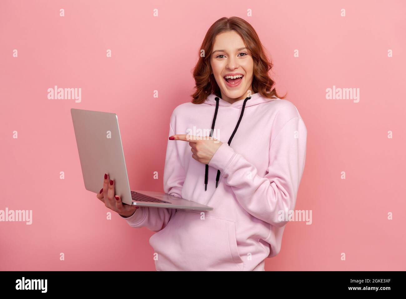 Porträt eines optimistischen, lockigen Teenagers im Kapuzenpullover, das lächelt und mit dem Finger auf das Notebook zeigt und ein gutes Ergebnis genießt. Innenaufnahmen im Studio iso Stockfoto