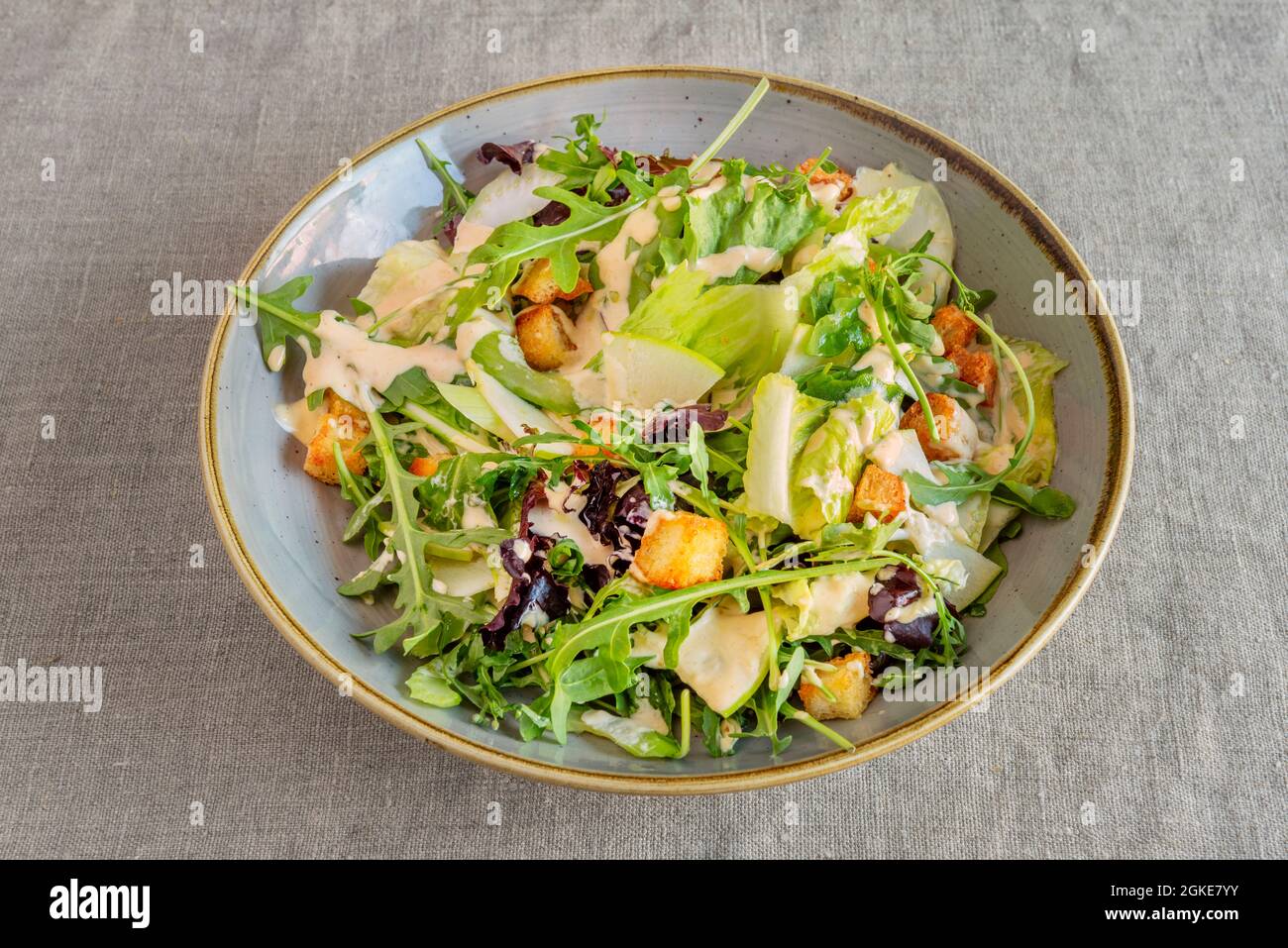 Schale grüner Salat mit Apfelscheiben, Käsescheiben, Croutons, Rucola und Salatsprossen mit Mayonnaise-Sauce auf grauer Leinentischdecke Stockfoto