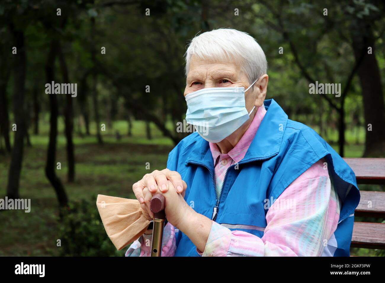 Ältere Frau in Schutzmaske sitzt mit Gehstock auf einer Bank im Park. Sicherheit während einer Coronavirus-Pandemie, Leben im Ruhestand Stockfoto