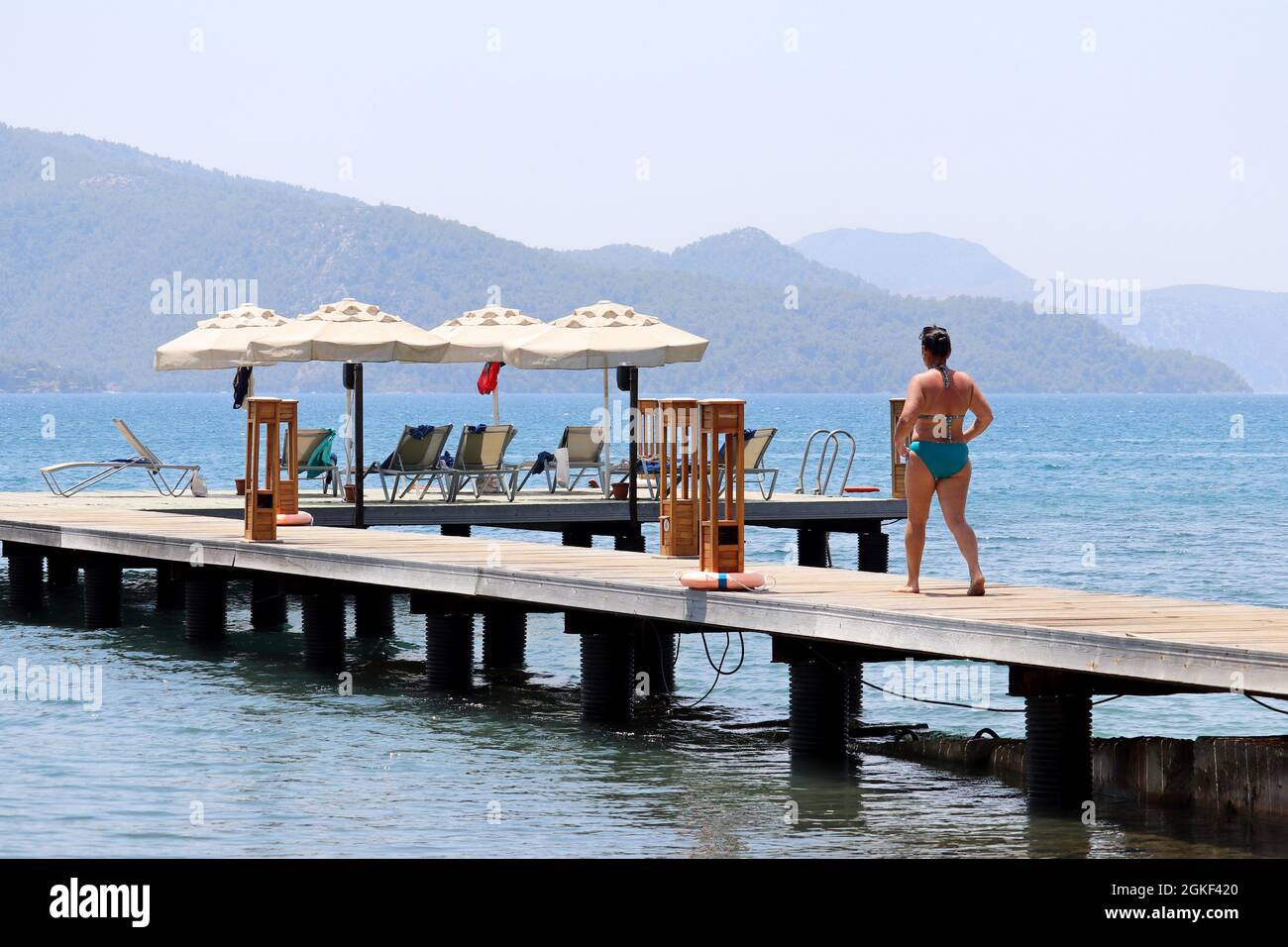 Strandurlaub im Seebad, Frau im Badeanzug, die mit Liegestühlen und Sonnenschirmen auf dem Pier spazierengeht. Landschaftlich schöner Blick auf die grüne Berginsel im Nebel Stockfoto