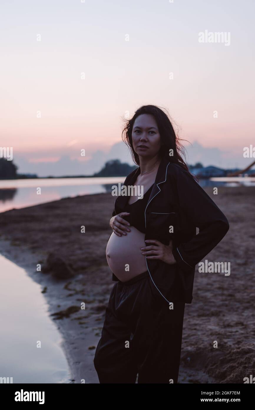 Porträt einer Schwangeren am Strand. Romantisches Foto von attraktiven jungen Brünette in unknöpfbaren dunklen Schlafanzug am Strand Blick auf Kamera und ihr Haar Stockfoto