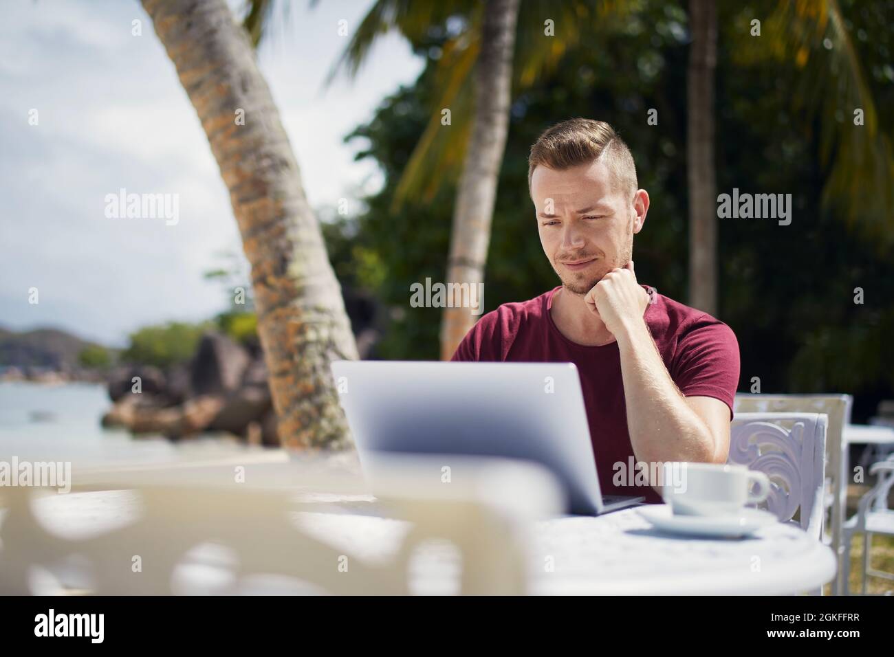 Junge Freiberuflerin, die an einem Laptop von einem tropischen Reiseziel aus arbeitet. Mann, der am Strand unter Palmen sitzt. Stockfoto
