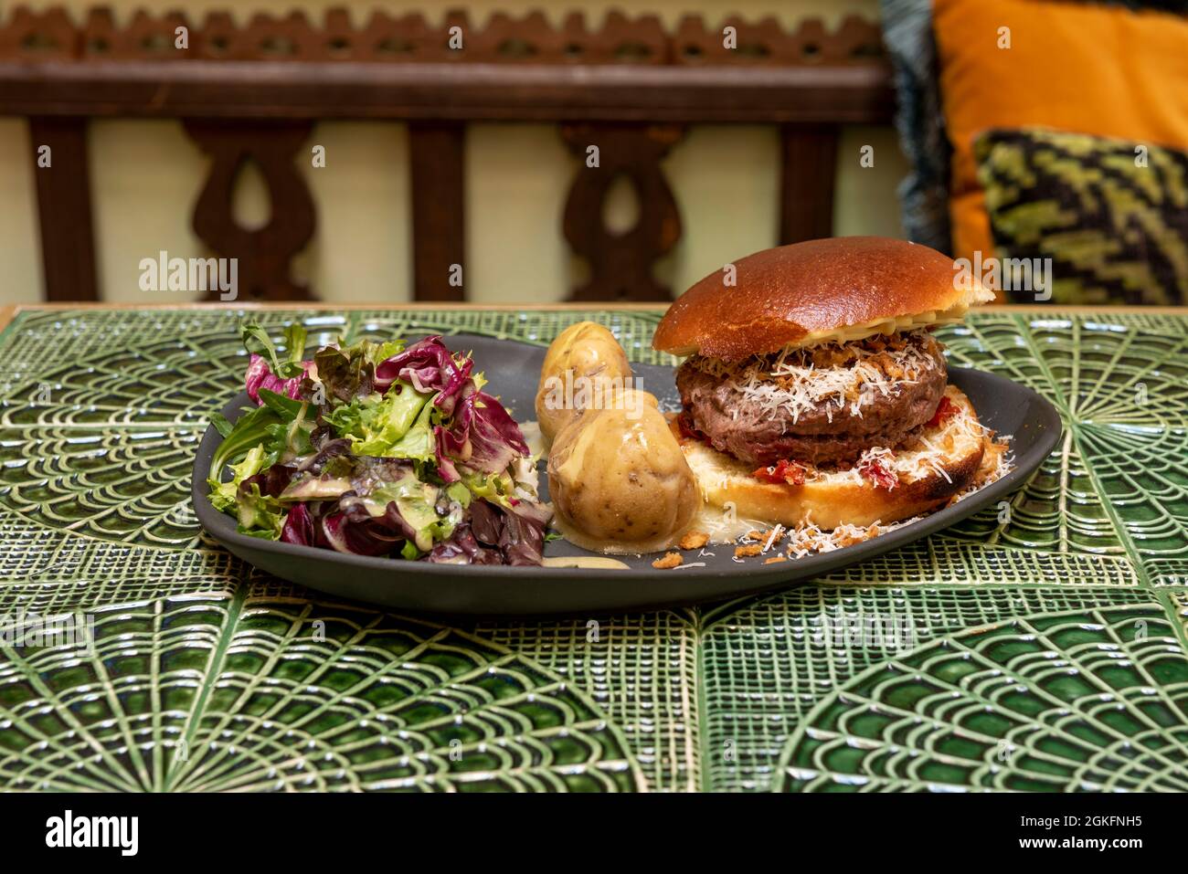 Menü eines großen Hamburgers mit einem Stück Rindfleisch, das mit geriebenem Käse überfüllt ist, mit einer Garnierung von ganzen gerösteten Kartoffeln und einem Salat mit verschiedenen Sprossen Stockfoto