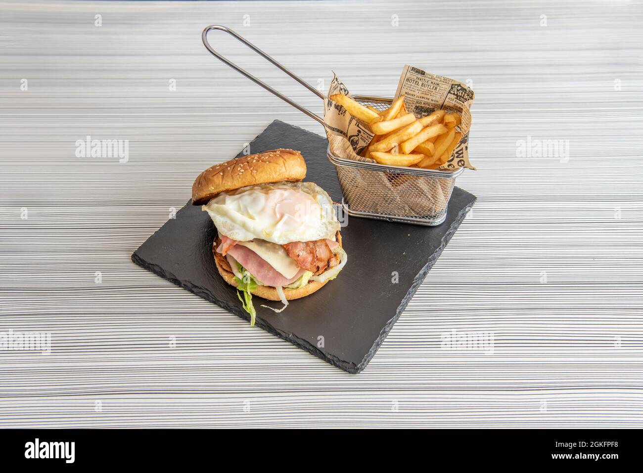 Köstlicher Rindfleisch-Burger mit Spiegelei, so dass sie versuchen, es zu essen, ohne es zu färben, begleitet von einem Korb mit pommes auf dem grauen Tisch Stockfoto