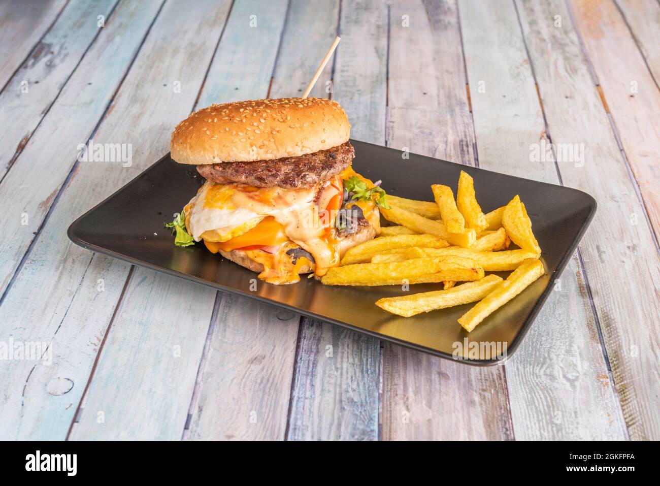 Rindfleisch-Burger mit gebratenem Käse, der auf Käse und Tomaten und Salatsprossen tropft, garniert mit Pommes auf schwarzem Tablett Stockfoto