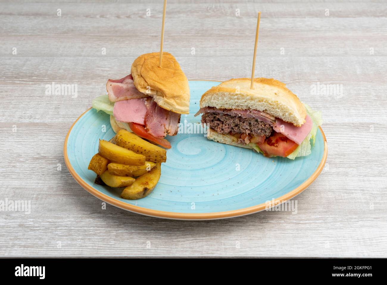 Rindfleisch-Burger mit Speck und Schinken, Tomaten und Salat in zwei Hälften geschnitten mit Pommes frites auf einem blauen Teller Stockfoto
