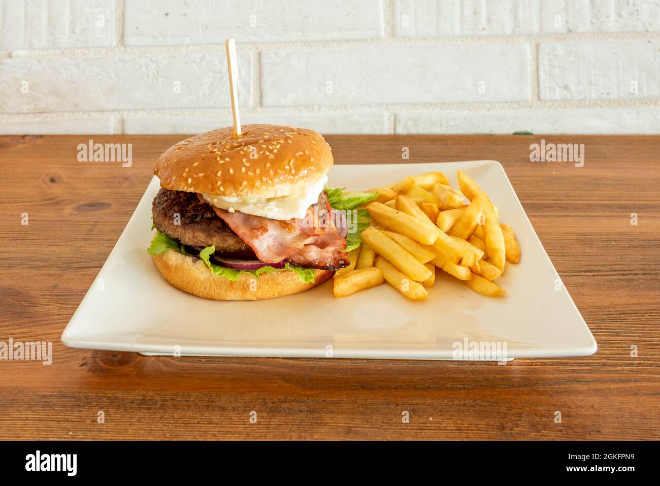 Rinderburger mit Speck, roter Zwiebel, Eisbergsalat und Spiegelei, garniert mit Pommes auf einem weißen quadratischen Teller Stockfoto