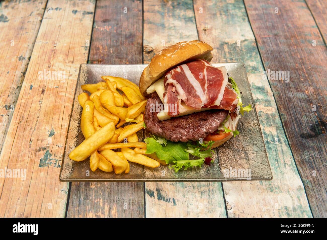 Rindfleisch-Burger mit Käse und Serrano-Schinken mit Salat mit einer Portion Pommes auf einer transparenten Glasplatte Stockfoto