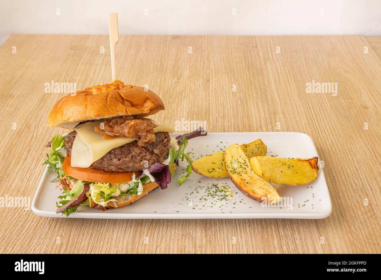 Rindfleisch-Burger mit Käse und karamellisierten Zwiebeln und einigen Kartoffelkeilen garnieren auf einem Eichenholztisch Stockfoto