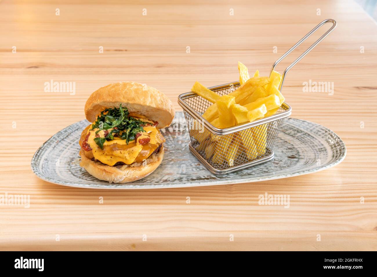 Spektakulärer Rindfleischburger mit viel Käse, gebratener Rucola und italienischer dehydrierter Tomate mit einer Portion Pommes als Beilage auf einem Holztisch Stockfoto