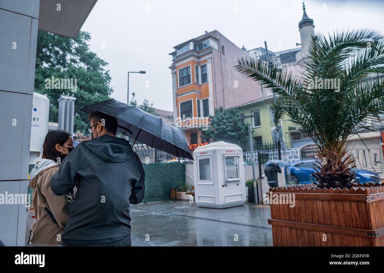 Beyoglu, Istanbul, Türkei - 06.18.2021: Männer- und Frauenfreunde mit schwarzem Regenschirm warten in der Nähe eines Gebäudes, um sich vor heftigem Regen draußen in Taken zu schützen Stockfoto