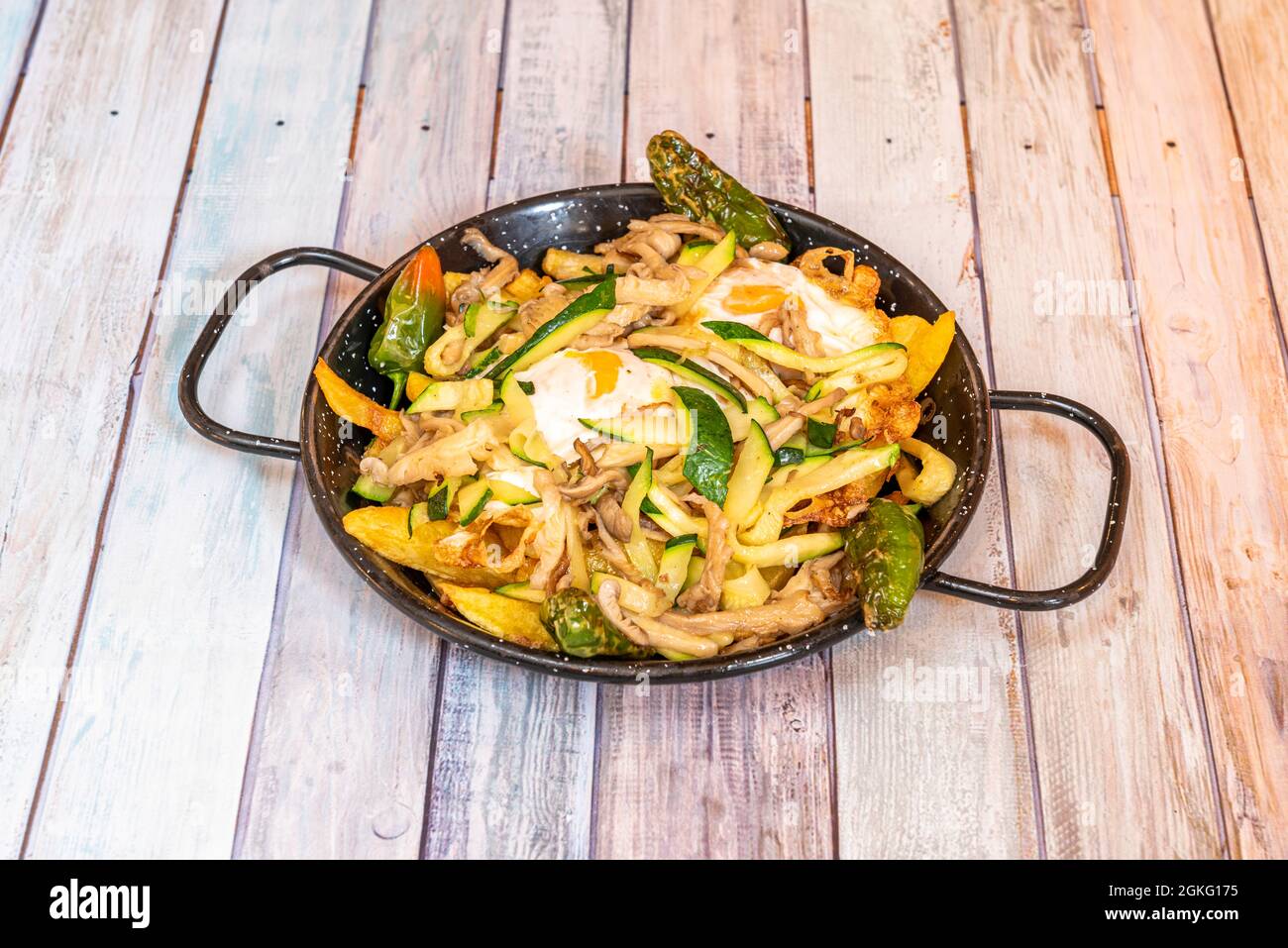 Rühreier mit Gemüse, Zucchini, Paprika, Pilzen und hausgemachten Pommes in schwarz emaillierter Paella-Pfanne auf Holztisch Stockfoto