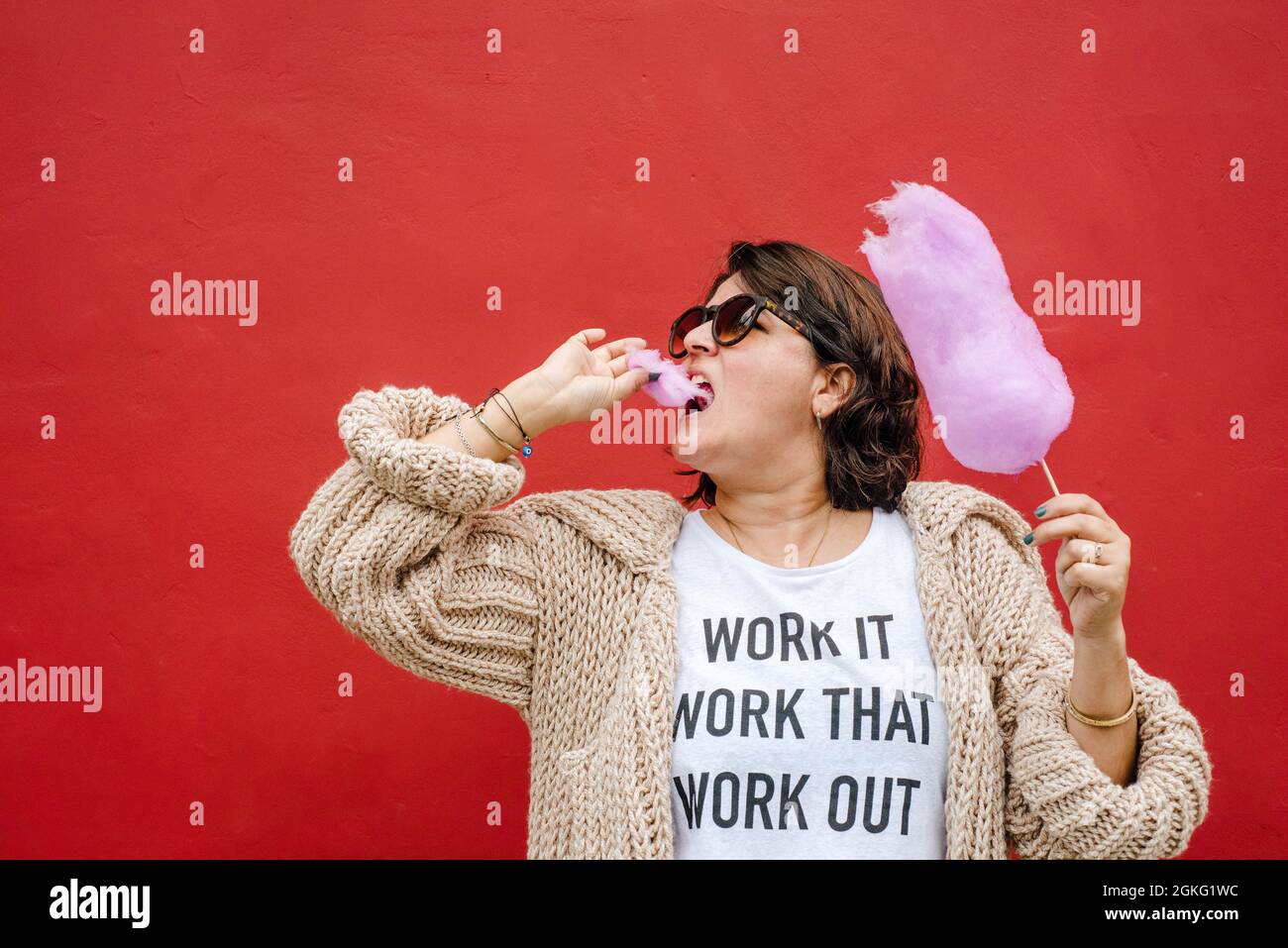 Eine Frau legt ihr ein Stück Zuckerwatte in den Mund. Sie tragen dunkle Gläser, Mantel auf rotem Hintergrund. Urbanes Bild Kopierbereich Stockfoto