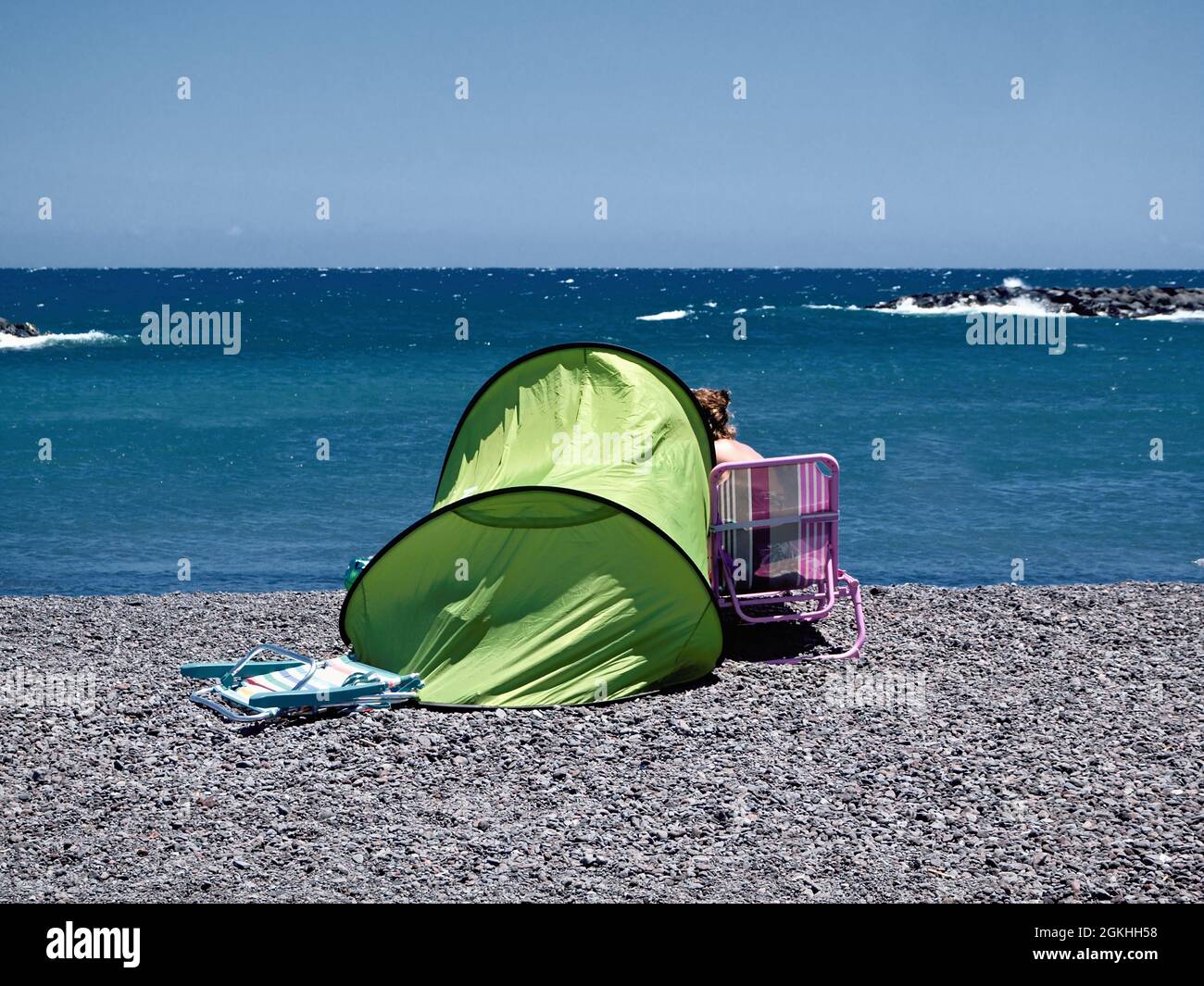 Ein kleines faßförmiges Sonnenschutzzelt am Kiesstrand vor dem Atlantik, daneben ein Campingstuhl, auf dem eine Frau mit Blick auf den Atlantik sitzt Stockfoto