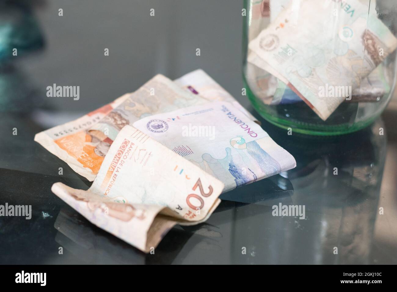 Kolumbianische Banknoten auf einem Glastisch, im Hintergrund ein Glas mit Geld, Wirtschaft, Bankrott, persönlichen Ersparnissen. Konzept von Wirtschaft und Armut Stockfoto